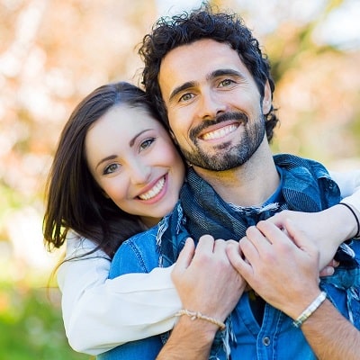 Smiling, dark-haired girl hugs bearded man from behind happy with their cosmetic dentistry treatment.